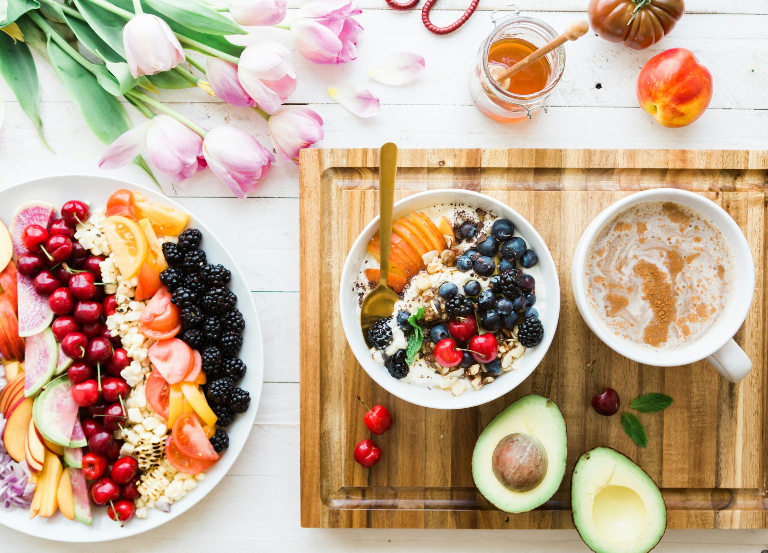 bowl of yogurt, plate of fruit, cup of coffee and sliced avocado on a wooden cutting board on a white background
