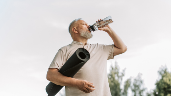 An older man drinks water from a plastic bottle and holds a black yoga mat